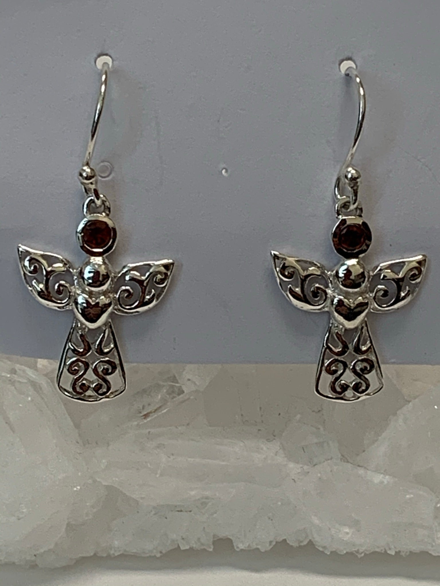 Close-up view. Small, round, faceted garnet gemstones are outlined in sterling silver and set above an angel's wings and dress (the garnet is in place of the angel's head/face). The angel's body and wings are filled with swirls of silver and there is a solid silver heart on the body, beneath the wings. This is a small and delicate pair of earrings. They are lightweight, have wires, not posts and are approximately 1½" long.