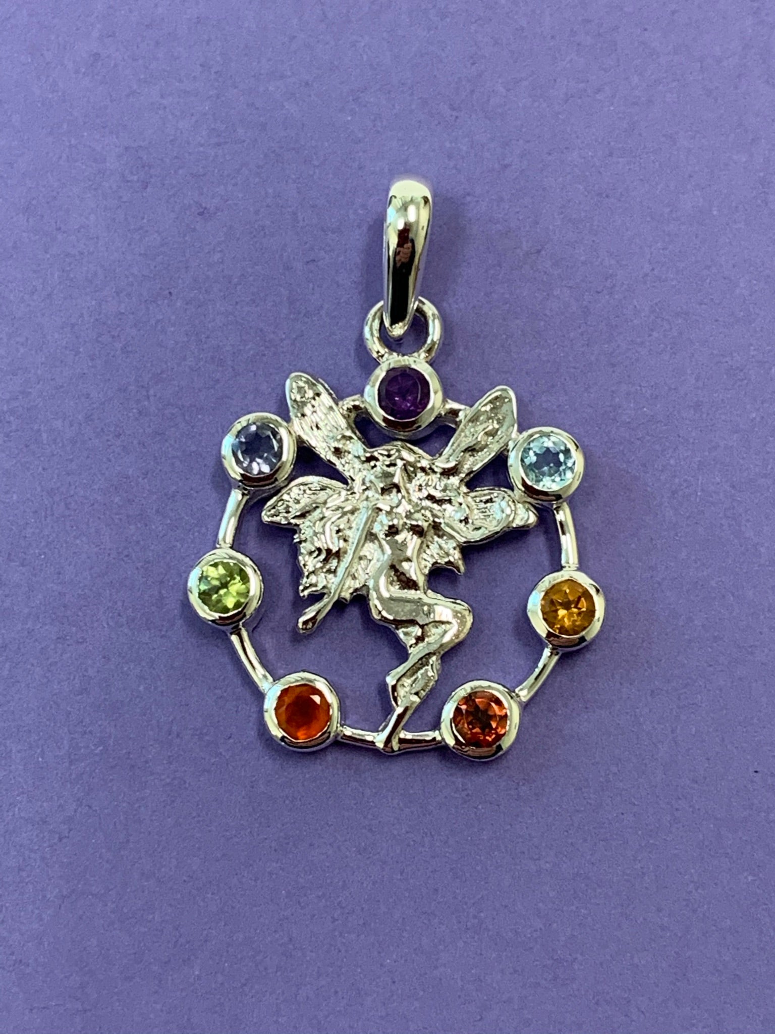 Another view of the Chakra fairy pendant. Small faceted stones represent each of the chakras: amethyst/crown, iolite/third eye, blue topaz/throat, peridot/heart, citrine/solar plexus, carnelian/sacral (or naval), garnet/root (or base). These small, faceted stones are set around an open sterling silver circle. A sterling silver fairy extends through the center of the circle. Her wings reach a bit beyond the top of the circle and her feet touch the bottom. Pendant is approximately 1".