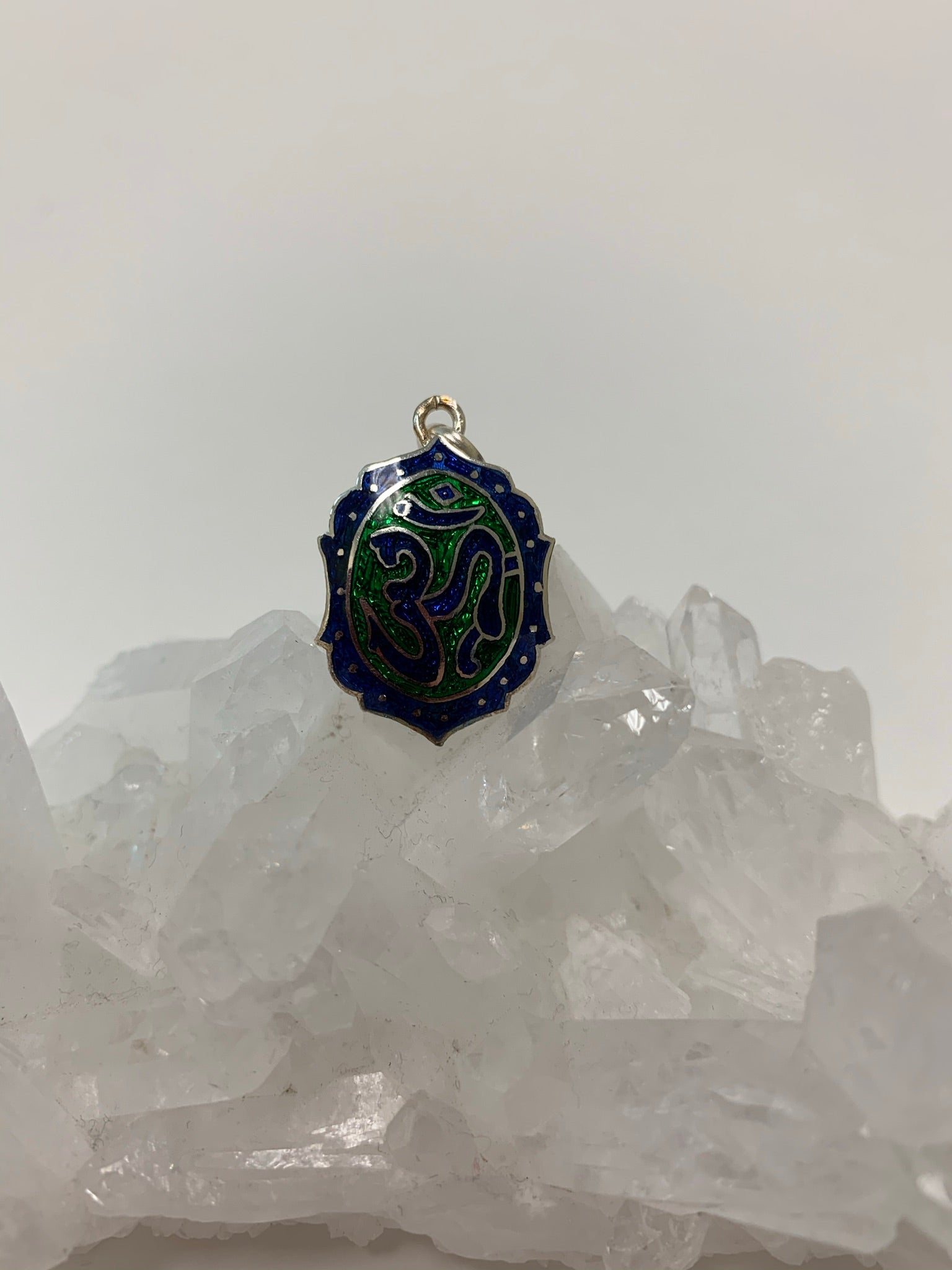 An additional view of the Om enamel pendant. It is oval-ish with the Om symbol in blue set within a green oval background. The Om oval is surrounded by a stylized oval-ish shape that is the same color blue as the om symbol. Around that edge. within the blue color, are tiny silver circles. The whole enamel pendant is set on sterling silver and has a sterling silver bail. This is a pendant only - no necklace chain included. It is approximately 1¼" long.