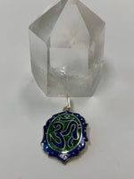 Load image into Gallery viewer, Om enamel pendant. It is oval-ish with the Om symbol in blue set within a green oval background. The Om oval is surrounded by a stylized oval-ish shape that is the same color blue as the om symbol.  Around that edge. within the blue color, are tiny silver circles. The whole enamel pendant is set on sterling silver and has a sterling silver bail. This is a pendant only - no necklace chain included. It is approximately 1¼&quot; long.
