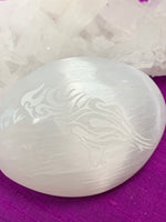 Load image into Gallery viewer, Close-up view of etched raven in smooth, polished selenite palm stone. Selenite ranges from translucent to milky white and is the perfect size to fit in your palm. Average weight is 4.2 oz and it is approximately 3&quot;x2.5&quot;. This selenite is ethically sourced and workers receive living wages.
