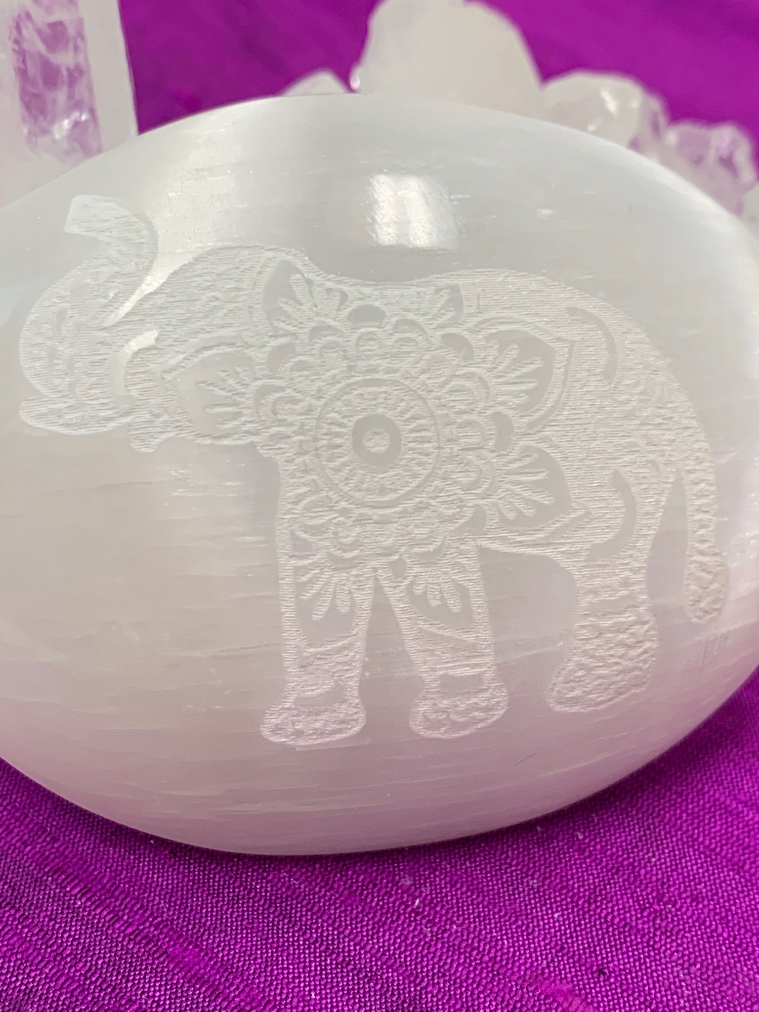 Close-up view of the ornately designed elephant etched on a smooth, polished selenite palm stone. The selenite stone can range from translucent to milky white in color. The weight of the stone varies, but the average weight is 4.2 oz and the average size is 3"x2.5", the perfect size to hold in the palm of your hand. These selenite stones are handmade and ethically sourced - workers are paid living wages.