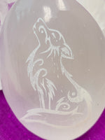 Load image into Gallery viewer, Close-up view of the wolf etched on smooth, polished white selenite palm stone. The selenite ranges from translucent to milky white in color. Wolf is intuitive, the teacher bringing knowledge back to the pack, is loyal and family oriented but also has an individualistic instinct. Weight varies, but the average is 4.2 oz and the size is approximately 3&quot;x2.5&quot;. These selenite stones are handcrafted and etched with lasers and by hand. Selenite is sourced ethically and workers are paid living wages.
