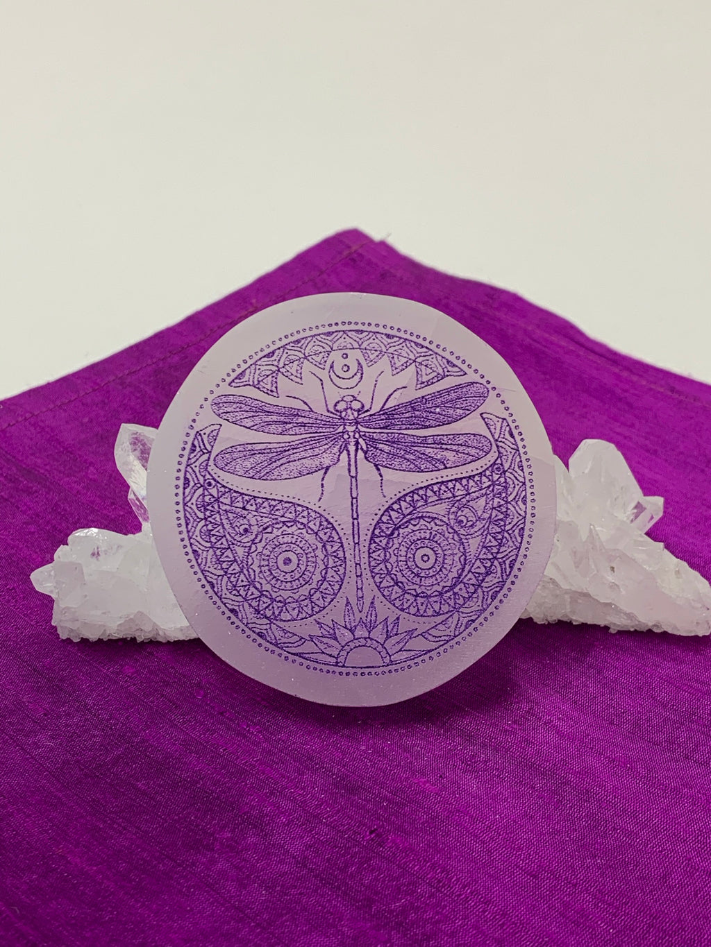 Beautiful 3" ornate dragonfly with flowing and detailed designs surrounding it is etched on smooth white selenite with the etching painted in purple. Dragonfly can represent emergence, dispelling of illusion and transformation. The selenite ranges from translucent to milky white in color. Weight varies, but the average is 4.9 oz. Size is approximately 3 inches in diameter and is approximately ½" thick. The selenite is ethically sourced and workers are paid living wages.