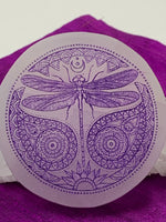 Load image into Gallery viewer, Close-up view of the beautiful 3&quot; ornate dragonfly with flowing and detailed designs surrounding it is etched on smooth white selenite with the etching painted in purple. Dragonfly can represent emergence, dispelling of illusion and transformation. The selenite ranges from translucent to milky white in color. Weight varies, but the average is 4.9 oz. Size is approximately 3 inches in diameter and is approximately ½&quot; thick. The selenite is ethically sourced and workers are paid living wages.
