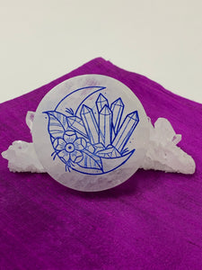 Side-facing crescent moon with crystal cluster and flower etched on white selenite plate (used to charge your crystals and jewelry). Etching is painted blue. The selenite ranges from translucent to milky white in color. Size varies, but average weight is 4.9 oz, size is approximately 3"x2.5" and approximately ½" thick. This selenite is ethically sourced and workers are paid living wages. 