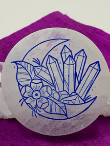 Close-up view of side-facing crescent moon with crystal cluster and flower etched on white selenite plate (used to charge your crystals and jewelry). Etching is painted blue. The selenite ranges from translucent to milky white in color. Size varies, but average weight is 4.9 oz, size is approximately 3"x2.5" and approximately ½" thick. This selenite is ethically sourced and workers are paid living wages.