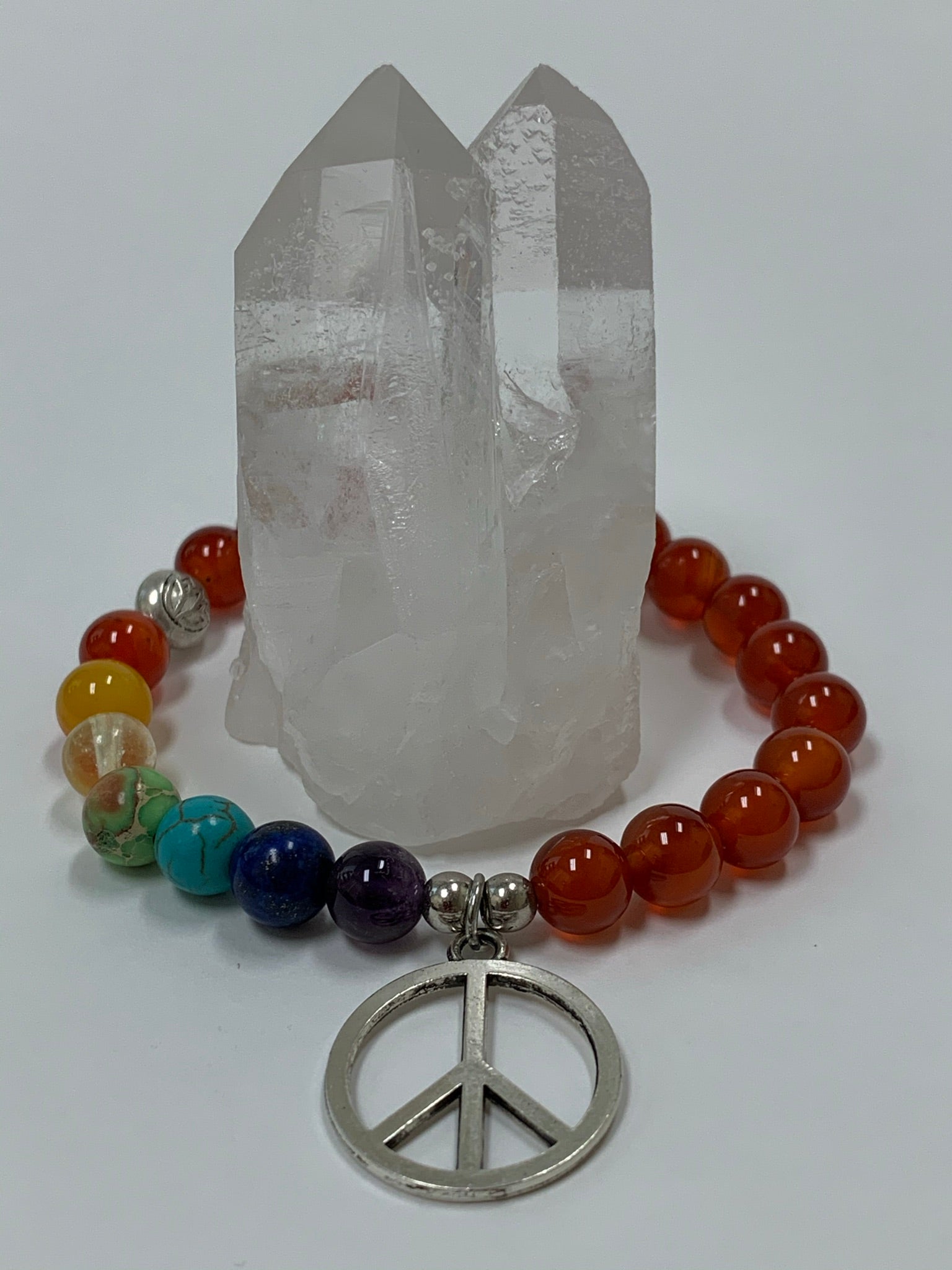 Close-up of the carnelian gemstone power bracelet, accented by 7 different gemstones, representing the 7 chakras and a silver-colored peace sign charm and 3 additional silver-colored beads (2 of which are plain and one has a lotus on it). These additional silver colored beads are not sterling silver.