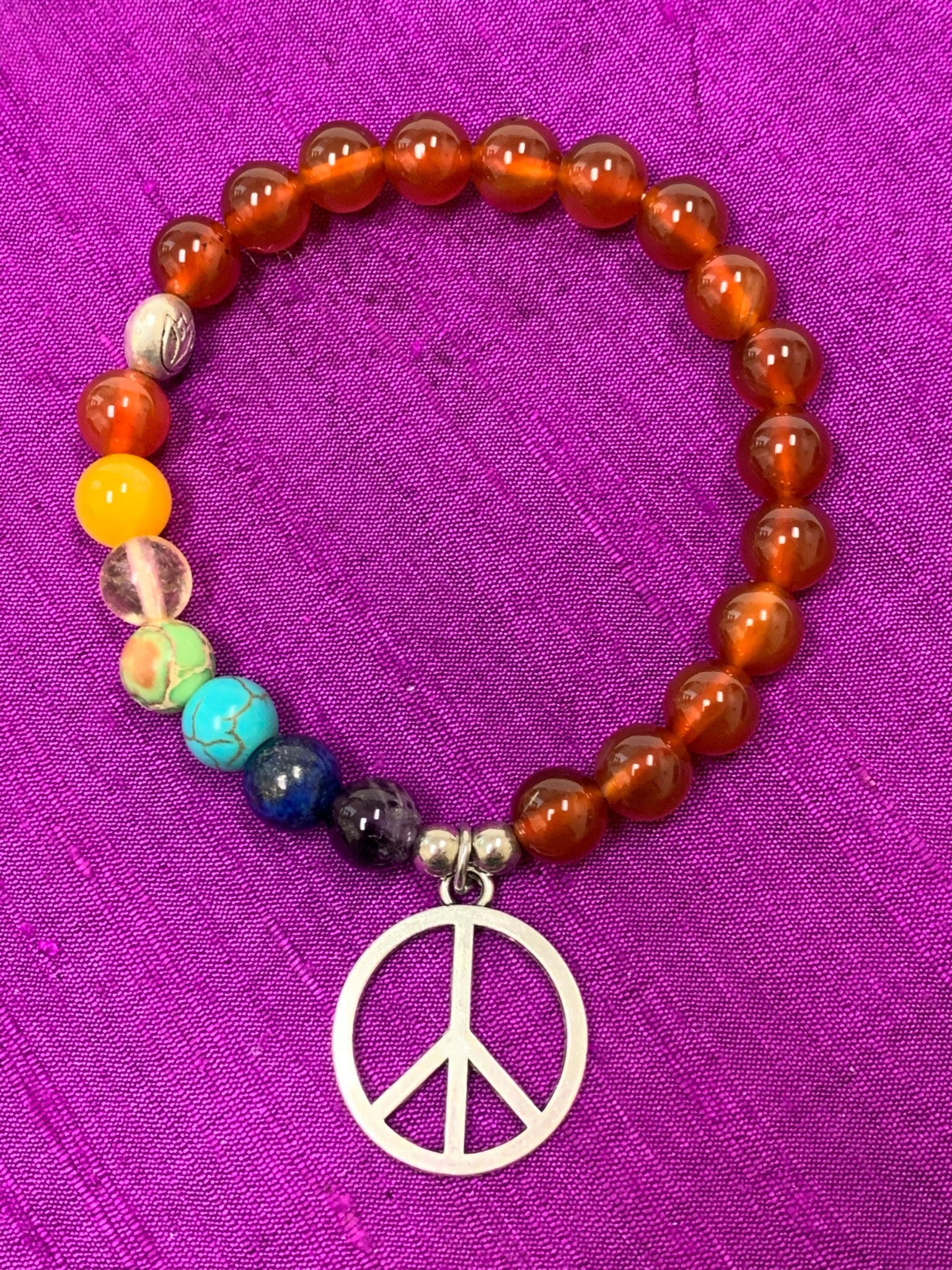 Second Close-up of the carnelian gemstone power bracelet, accented by 7 different gemstones, representing the 7 chakras and a silver-colored peace sign charm and 3 additional silver-colored beads (2 of which are plain and one has a lotus on it). These additional silver colored beads are not sterling silver.