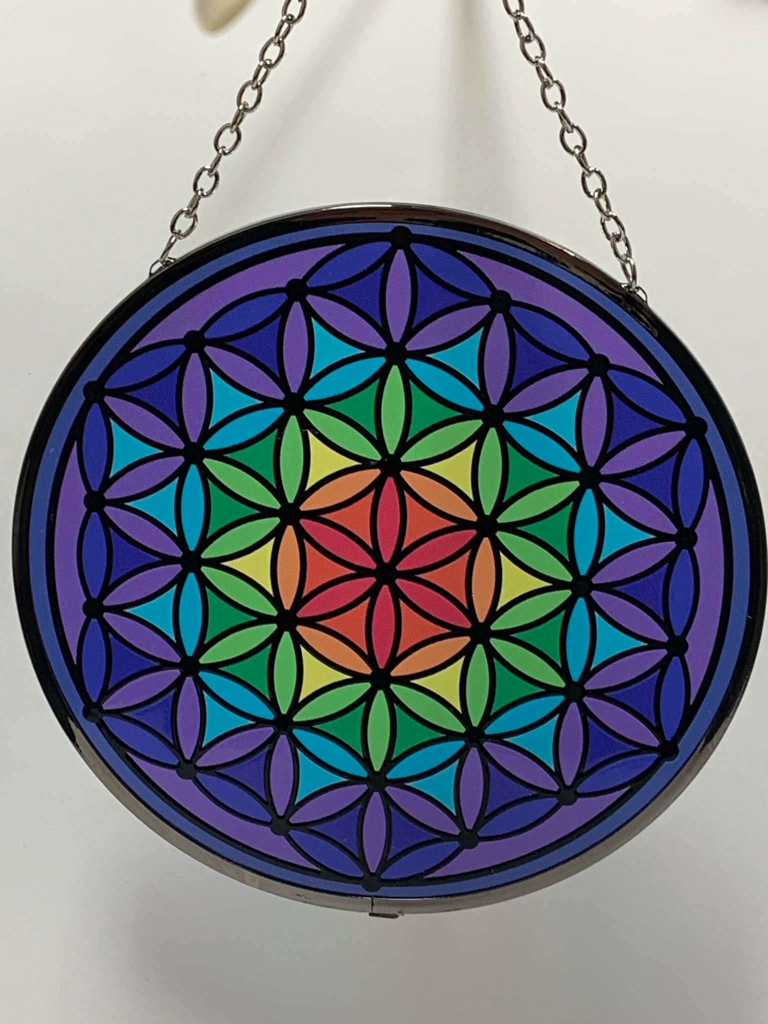 Close-up view. Round, glass flower of life suncatcher in the colors of the rainbow moving from the center (red) to the edge (purple). The already vibrant color is enhanced when the sun's rays stream through it. Beauty and spiritual meaning - nice combo! Comes with a chain for hanging and a suction cup, with a hook, to hold it.