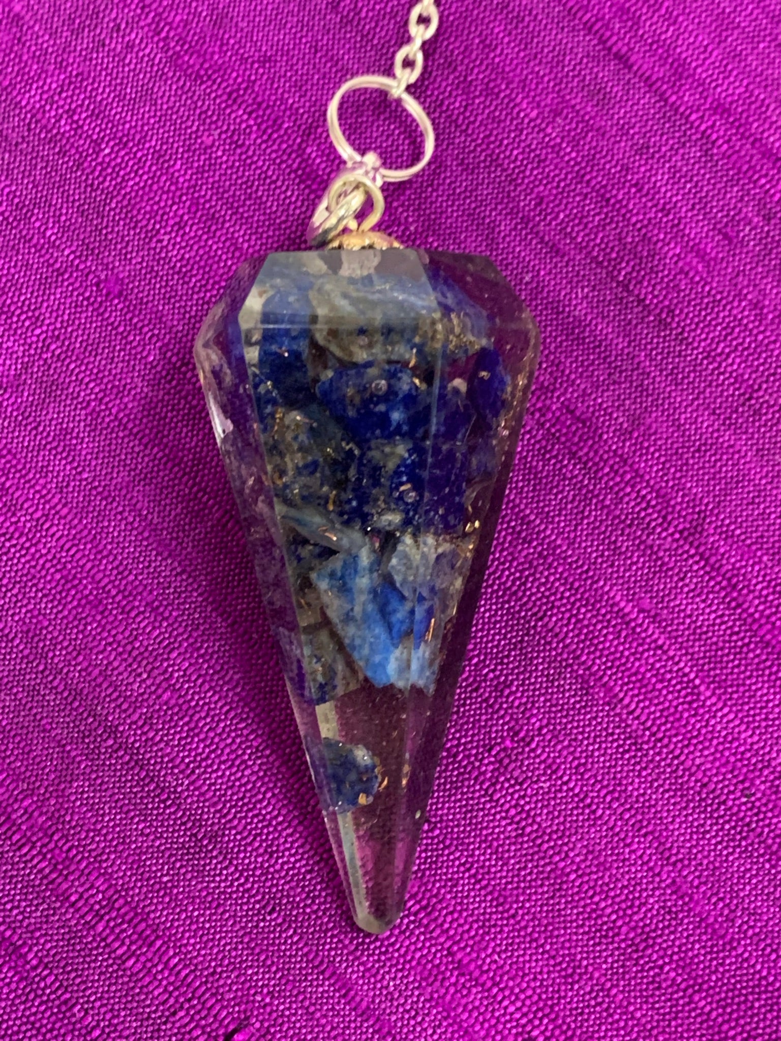 Close up view. Orgonite pendulum with lapis, accented with 7 chakra gemstone beads, on the its chain, 1 for each of the 7 major chakras. Also accented by a lapis gemstone heart at the end of the the pendulum's chain.