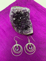 Load image into Gallery viewer, These amethyst rings of Venus earrings have three thin silver circles dangling from the ear wire. Each circle is bigger than the one before it and a faceted, tear drop amethyst hangs in the middle of the first and smallest circle. These earrings are pretty and fun! They are approximately 1¾&quot; long. 
