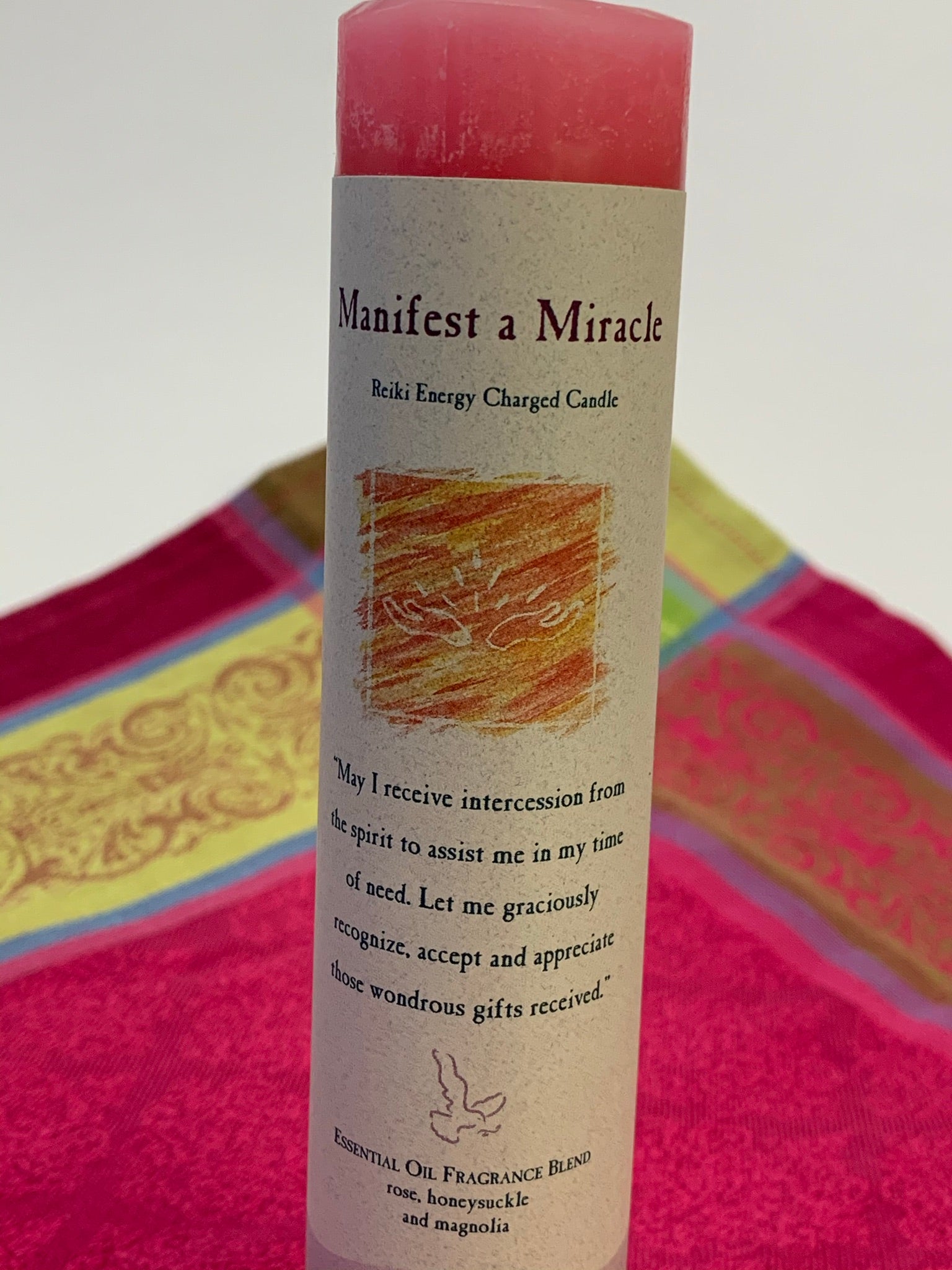 Close-up view of the Reiki-charged pillar candle for "manifesting a miracle." It is handcrafted and scented using essential oils. An affirmation/prayer is printed on the label, as well as a listing of the essential oils used (rose, honeysuckle & magnolia). It is approximately 7"x1½." Perfect for meditation, prayer, visualization or quiet time. This pink candle and its label are both visually appealing.