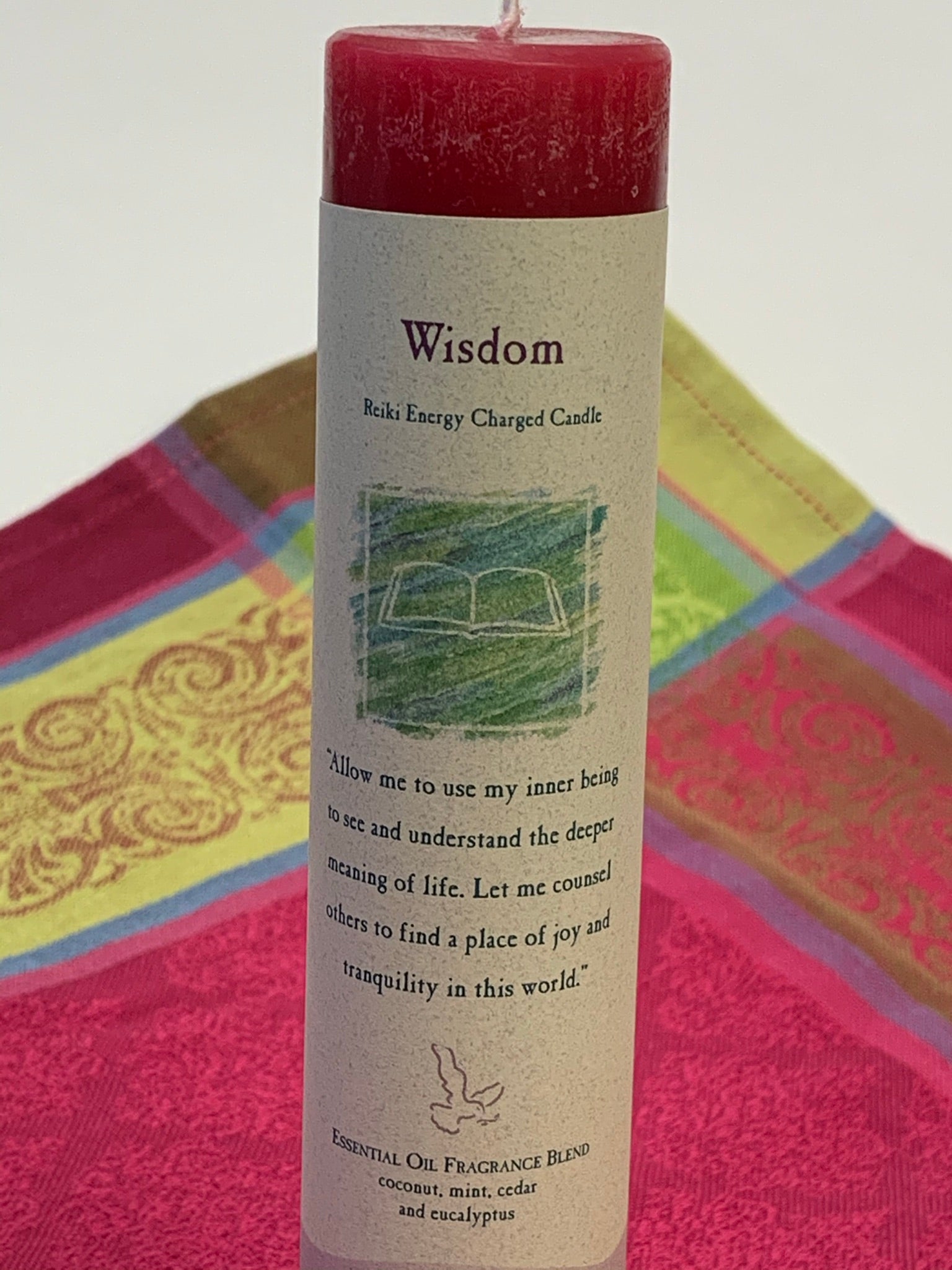Close-up view of the Reiki-charged pillar candle for "wisdom." It is handcrafted and scented using essential oils (coconut, mint, cedar & eucalyptus. An affirmation/prayer is printed on the label as well as a listing of the essential oils used. It is approximately 7"x1½". Perfect for meditation, prayer, visualization or quiet time. This red candle and its label are both visually appealing.