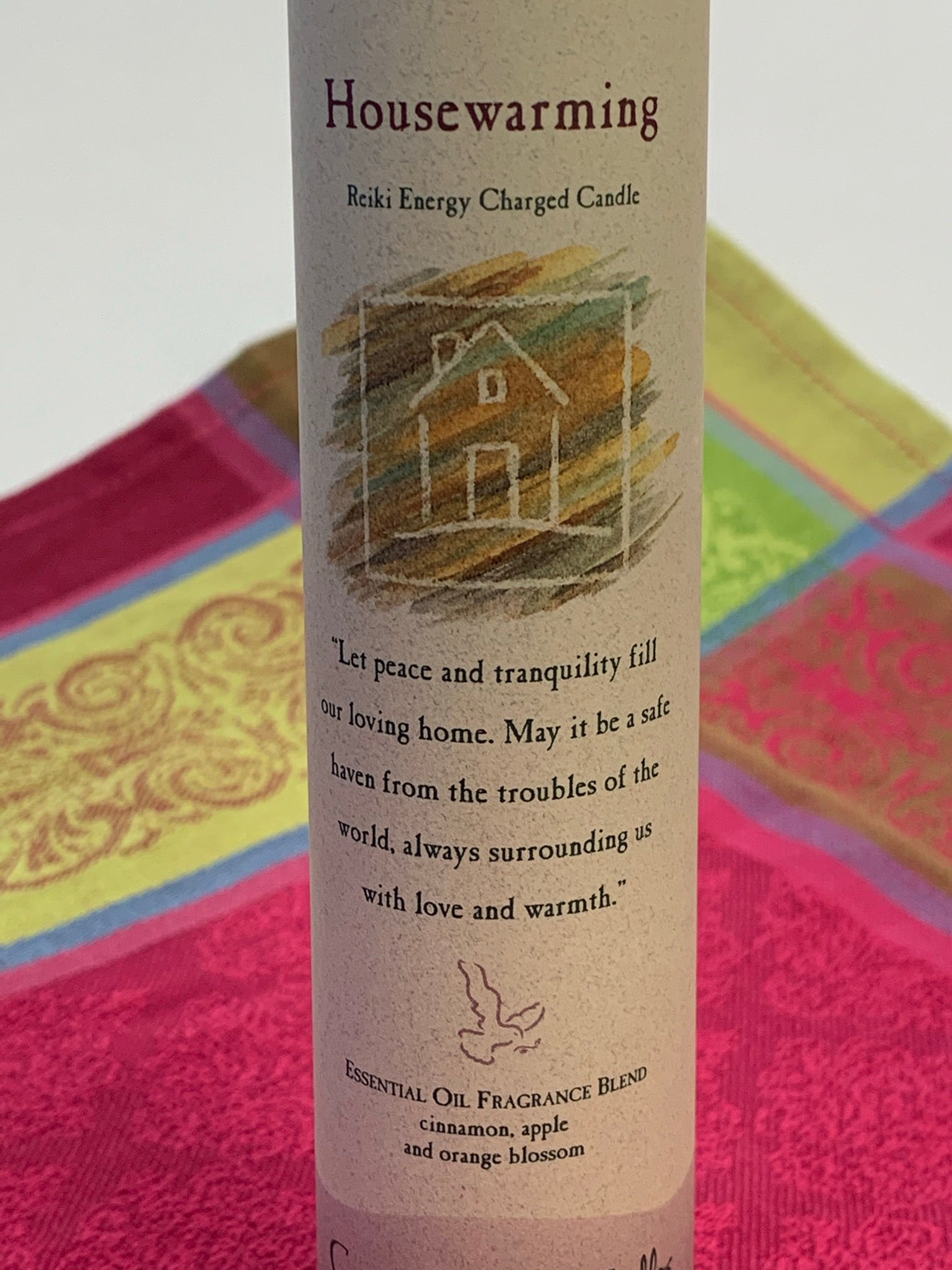 Close-up view of the Reiki-charged pillar candle for "Housewarming" energy - bringing love, peace and warmth to any home. It is handcrafted and scented using essential oils. An affirmation/prayer is printed on the label, a well as a listing of the essential oils used (cinnamon, apple and orange blossom). It is approximately 7"x2½". Perfect for meditation, visualization or quiet time. This mahogany brown candle and its label are both visually appealing.