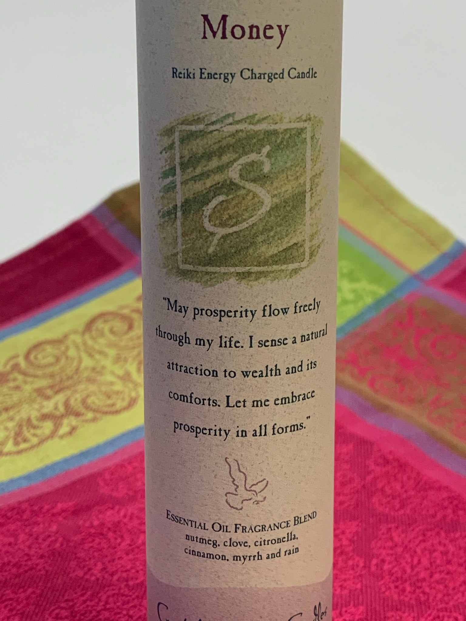 Close-up view of the Reiki-charged pillar candle energized for manifesting "money." It is handcrafted and scented using essential oils. An affirmation/prayer is printed on the label, as well as a listing of the essential oils used (nutmeg, clove, citronella, cinnamon, myrrh, rain). It is approximately 7"x2½". Perfect for meditation, prayer, visualization or quiet time. This green candle and its label are both visually appealing.