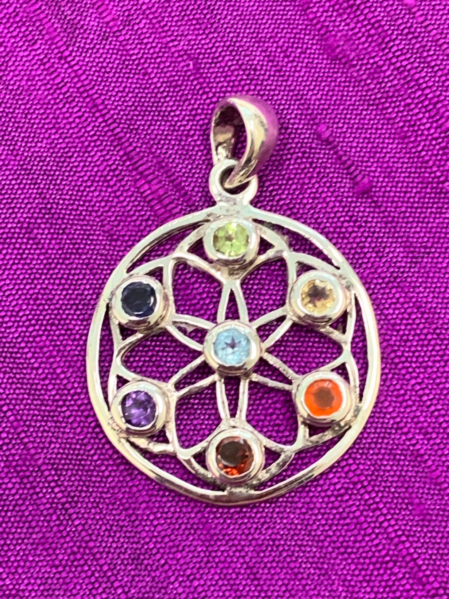 Close-up view. Beautiful chakra wheel pendant with seven small faceted stones around the wheel, each representing one of the 7 major chakras. Stones are set in sterling silver wheel. Chakras are energy centers in our bodies that process energy - coming into and flowing out of the body. The pendant is approximately 1¼".