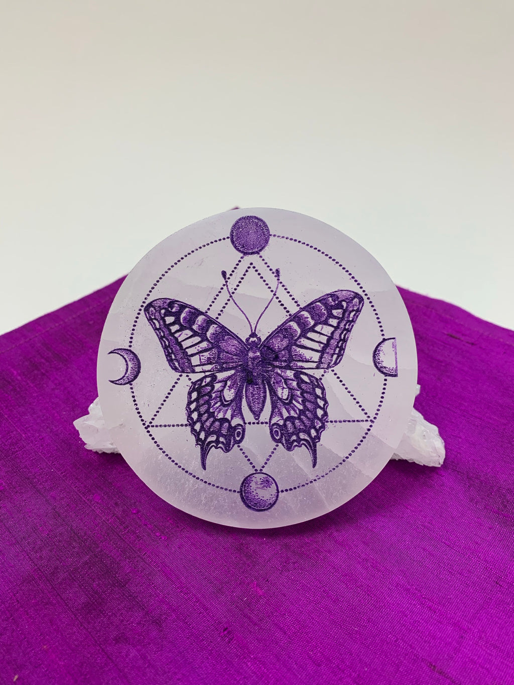 Gorgeous white selenite charging plate with a butterfly in the middle of the plate and moon phases (4) around her with circular and triangular dot designs. Butterfly and other designs are painted purple. The plate is approximately 4" in diameter and approximately ½" thick. It weighs about 9.1 ounces. Butterfly symbolizes transformation and the moon symbolizes feminine energy, intuition and emotions. Selenite plates are used for charging other crystals, gemstones and jewelry. 