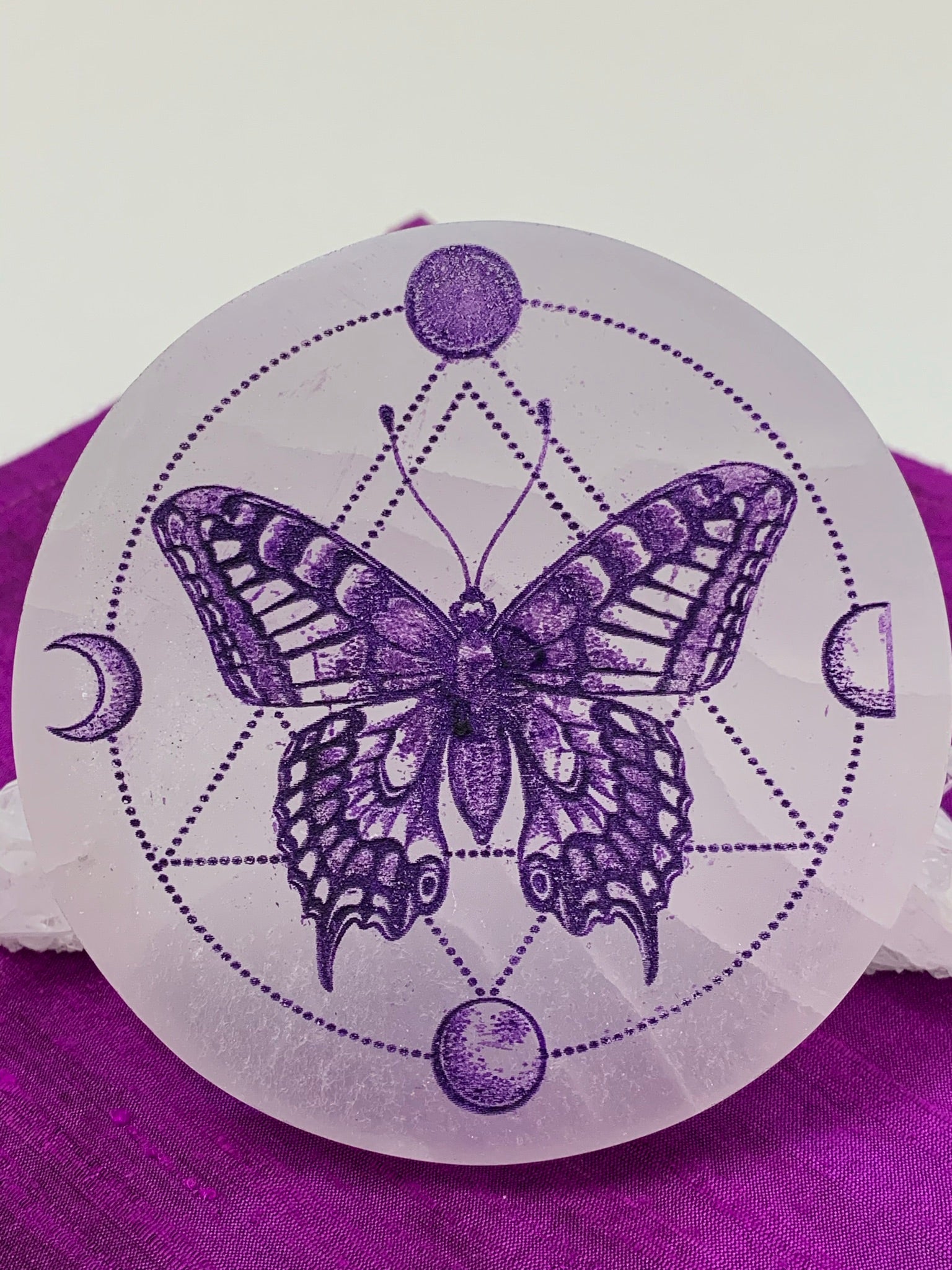 Close-up view. Gorgeous white selenite charging plate with a butterfly in the middle of the plate and moon phases (4) around her with circular and triangular dot designs. Butterfly and other designs are painted purple. The plate is approximately 4" in diameter and approximately ½" thick. It weighs about 9.1 ounces. Butterfly symbolizes transformation and the moon symbolizes feminine energy, intuition and emotions. Selenite plates are used for charging other crystals, gemstones and jewelry.