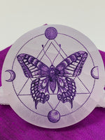 Load image into Gallery viewer, Close-up view. Gorgeous white selenite charging plate with a butterfly in the middle of the plate and moon phases (4) around her with circular and triangular dot designs. Butterfly and other designs are painted purple. The plate is approximately 4&quot; in diameter and approximately ½&quot; thick. It weighs about 9.1 ounces. Butterfly symbolizes transformation and the moon symbolizes feminine energy, intuition and emotions. Selenite plates are used for charging other crystals, gemstones and jewelry.
