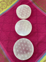 Load image into Gallery viewer, View of several selenite moons. Beautiful selenite full moon charging plate, etched with the flower of life, can be used for cleansing and charging your crystals and jewelry because it dispels negative and unwanted energies. Selenite also promotes clarity of mind, opens the crown chakra, helps to access angelic energies and guidance from the higher realms. It brings peace and is a great stone to use while meditating or connecting spiritually and more. Approx. 3&quot;. Cost is $10.
