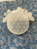 Load image into Gallery viewer, Alternate view. Beautiful selenite full moon charging plate, etched with the flower of life, can be used for cleansing and charging your crystals and jewelry because it dispels negative and unwanted energies. Selenite also promotes clarity of mind, opens the crown chakra, helps to access angelic energies and guidance from the higher realms. It brings peace and is a great stone to use while meditating or connecting spiritually and more. Approx. 3&quot;. Cost is $10.
