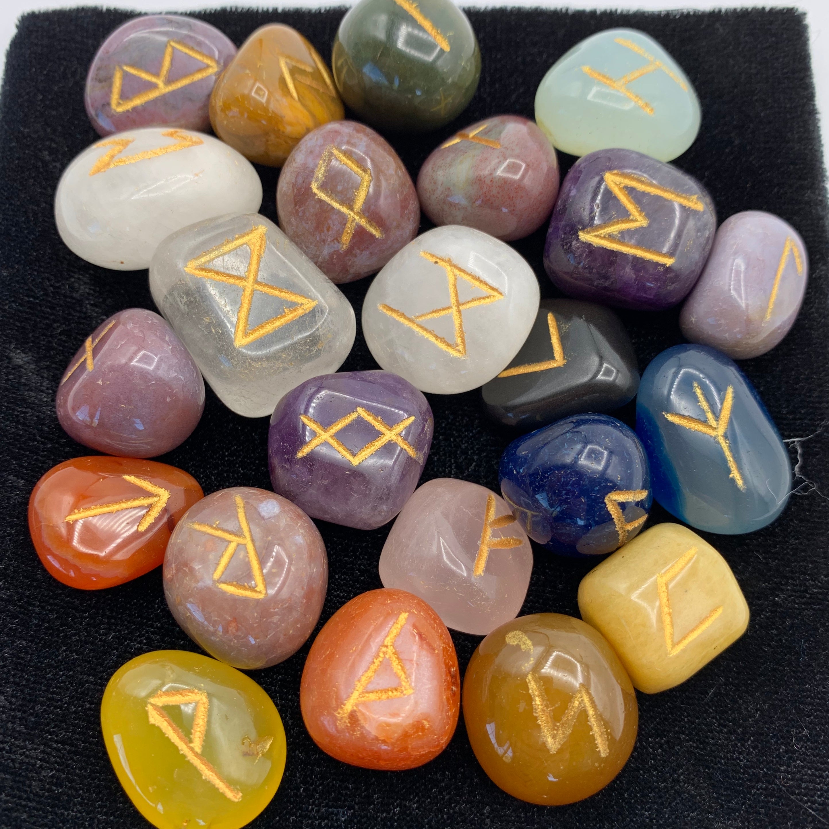 Close-up view of the mixed tumbled gemstone rune set with runic symbols on each stone in gold. Mixed tumbled stones means there will be a variety of gemstones, not just one type. There is no guarantee which stones will be included, but in this photo there are some amethyst, clear quartz, carnelian and more - very visually appealing. The runes come in a velvet bag with a paper sheet of rune meanings.