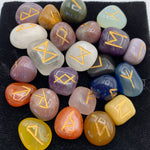 Load image into Gallery viewer, Close-up view of the mixed tumbled gemstone rune set with runic symbols on each stone in gold. Mixed tumbled stones means there will be a variety of gemstones, not just one type. There is no guarantee which stones will be included, but in this photo there are some amethyst, clear quartz, carnelian and more - very visually appealing. The runes come in a velvet bag with a paper sheet of rune meanings.
